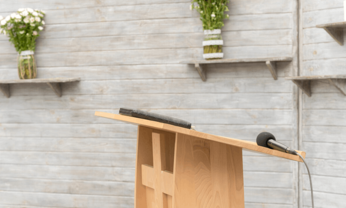 A wooden lectern with a microphone and book, set against a light timber wall with floral arrangements.